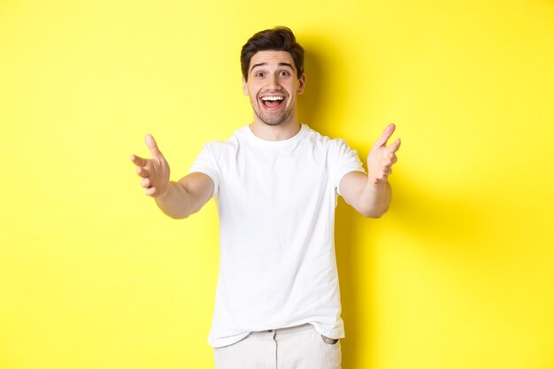 Excited handsome guy stretching hands forward, reaching for hug, receiving gift, standing over yellow background.