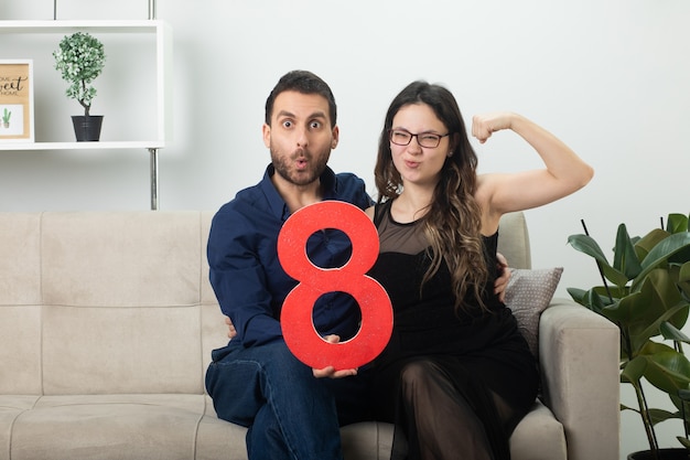 Excited handsome man holding red eight figure and pretty young woman in optical glasses tensing her biceps sitting on couch in living room on march international women's day