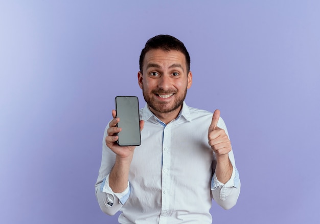 Excited handsome man holds phone thumbs up isolated on purple wall