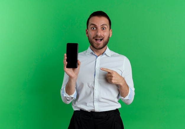 Excited handsome man holds and points at phone isolated on green wall