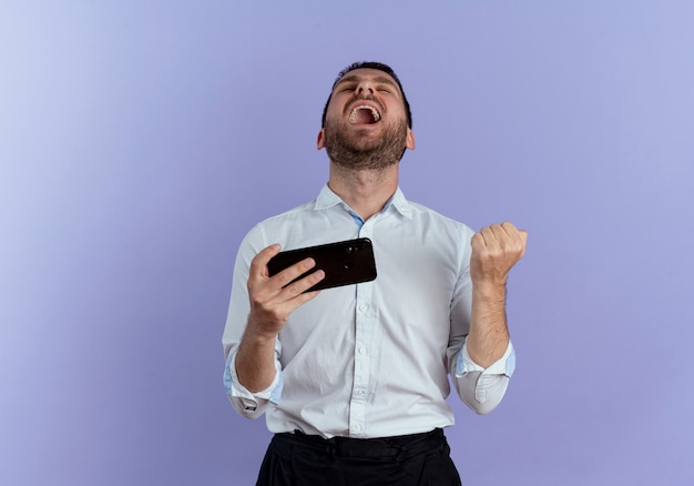 Free photo excited handsome man keeps fist and holds phone looking up isolated on purple wall