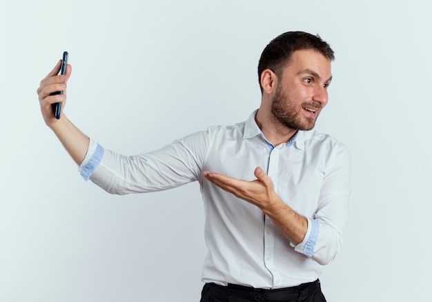 Excited handsome man looks at side holding and pointing at phone isolated on white wall