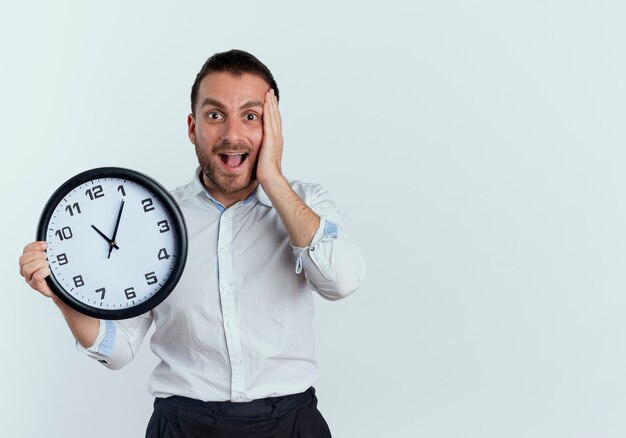 Excited handsome man puts hand on face holding clock isolated on white wall