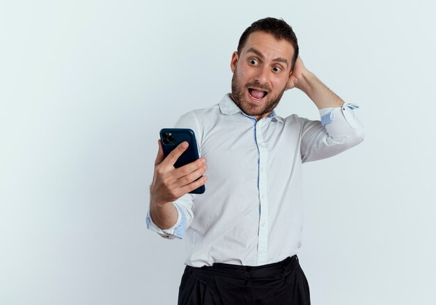 Excited handsome man puts hand on head behind looking at phone isolated on white wall