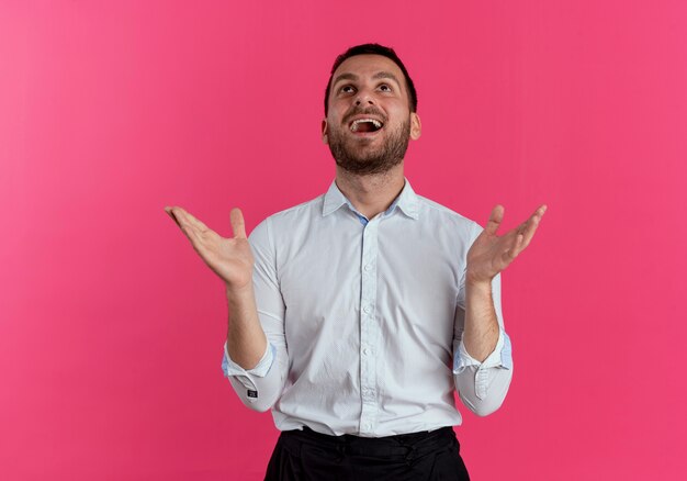 Excited handsome man raises hands looking up isolated on pink wall