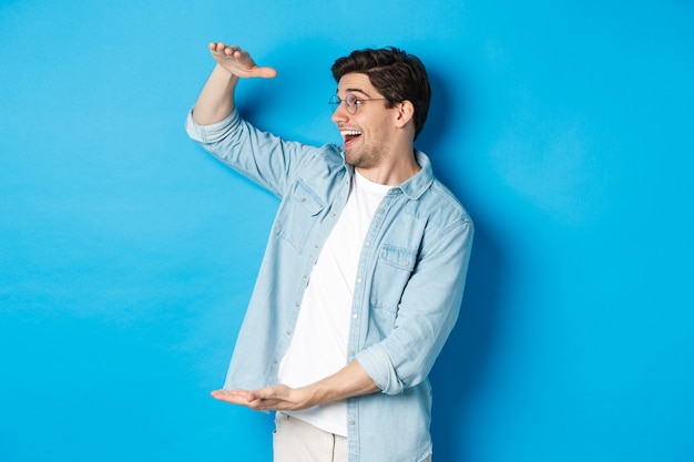 Excited handsome man showing big size object and looking amazed, standing over blue background