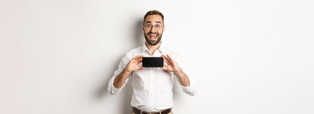 Excited handsome man showing mobile phone screen looking amazed standing over white background