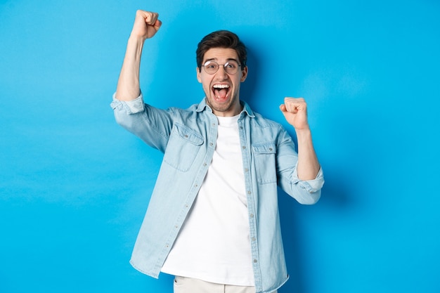 Excited handsome man triumphing, raising hands up and shouting for joy, celebrating victory, standing against blue background