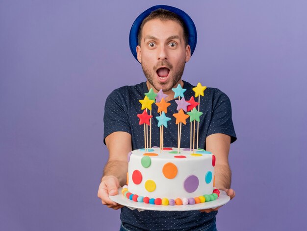 Excited handsome man wearing blue hat holds birthday cake looking at front isolated on purple wall