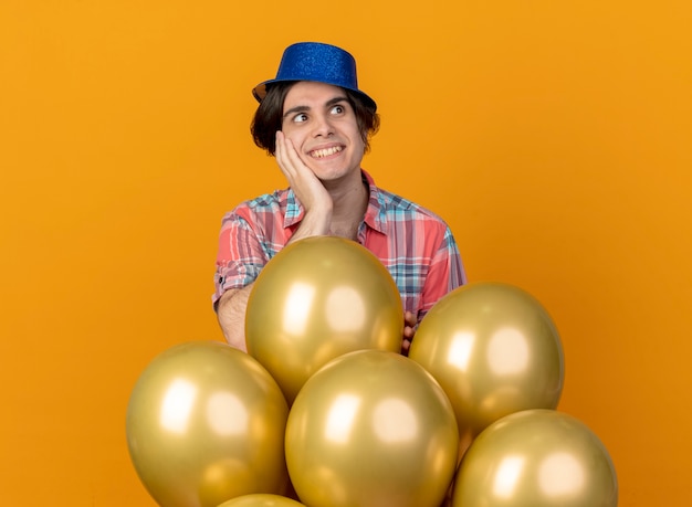 Excited handsome man wearing blue party hat puts hand on face and stands with helium balloons looking at side isolated on orange wall