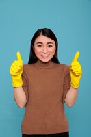 家事から興奮した幸せで陽気なメイドは親指を立て、青い背景に手の安全のために黄色の手袋を着用し、家の掃除のコンセプト、ポジティブなムードを持つ満足した空き地の女性