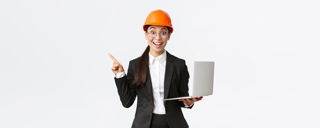Excited happy asian female engineer industrial woman in safety helmet and business suit showing pres