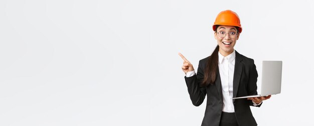 Взволнованная счастливая азиатская женщина-инженер, промышленная женщина в защитном шлеме и деловом костюме, показывающая презентацию, указывающая пальцем на график или диаграмму и держащая ноутбук, удивленно улыбаясь