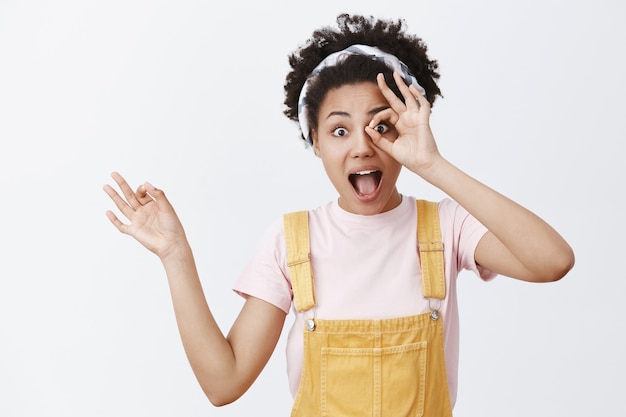 Excited and happy attractive african-american stylish female in yellow overalls over t-shirt, showing ok or zero gesture over eye and gazing thrilled, feeling joyful and amused