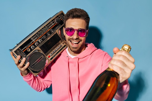 Excited happy bearded man in pink sunglasses and hoodie holds record player and champagne bottle on blue background