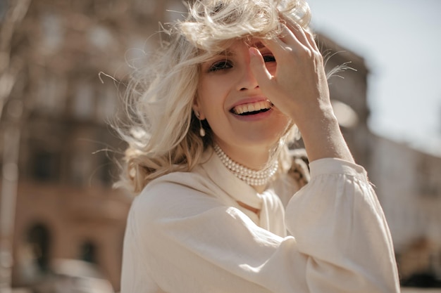 Бесплатное фото Возбужденная счастливая блондинка в жемчужном ожерелье abc белая блузка искренне улыбается, трогает волосы и позирует на улице