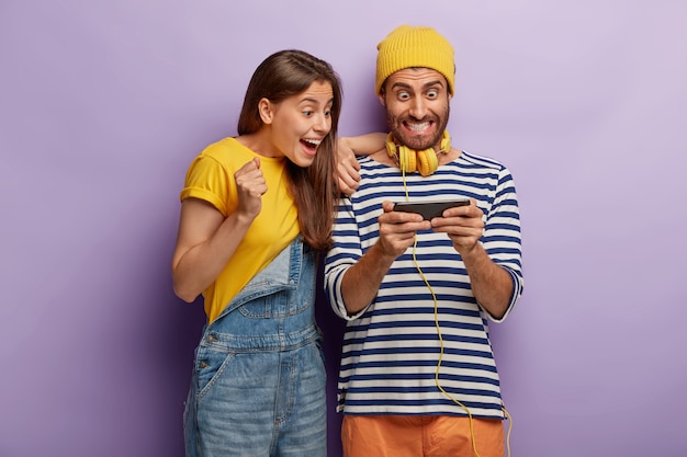 Excited happy couple use mobile phone for playing online games, look impressively at smartphone device, being obssessed with modern technologies, dressed in fashionable clothes. internet addiction