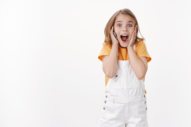 Excited happy emotive young girl with blond short hair wear summer overalls, touch cheek impressed, fascinated hear incredible awesome news, hold mobile phone, calling friend via smartphone