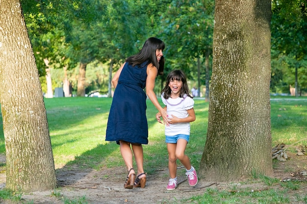 Взволнованная счастливая девушка играет в активные игры со своей мамой на открытом воздухе, стоит у деревьев в парке и смеется. Полная длина. Концепция семейного активного отдыха и досуга