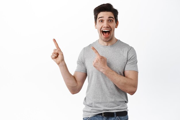 Excited happy guy points aside and laughs Joyful man shows advertisement on left side stands over white background