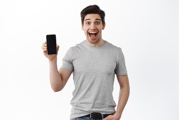 흥분된 행복한 남자는 흰색 배경에 서 있는 온라인 광고 스마트폰을 만드는 휴대전화 빈 화면을 보여줍니다.