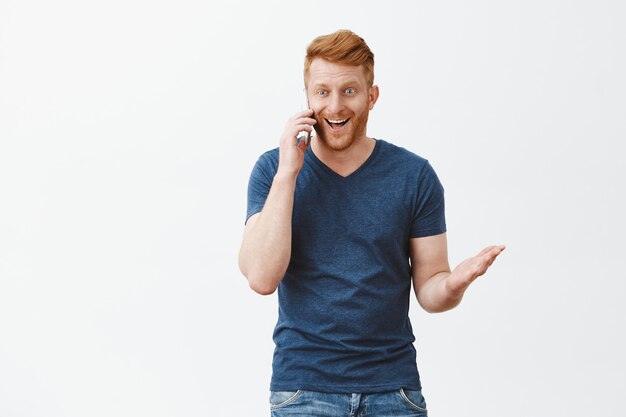 Excited and happy handsome redhead guy with brisle, gesturing with palm looking aside and smiling while talking on smartphone with thrill and joy