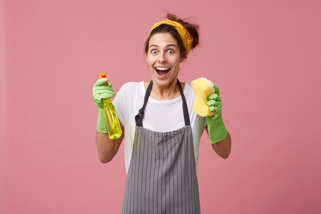 Excited happy housewife wearing yellow scarf on head and apron holding spray with sponge looking with widely opened mouth and eyes