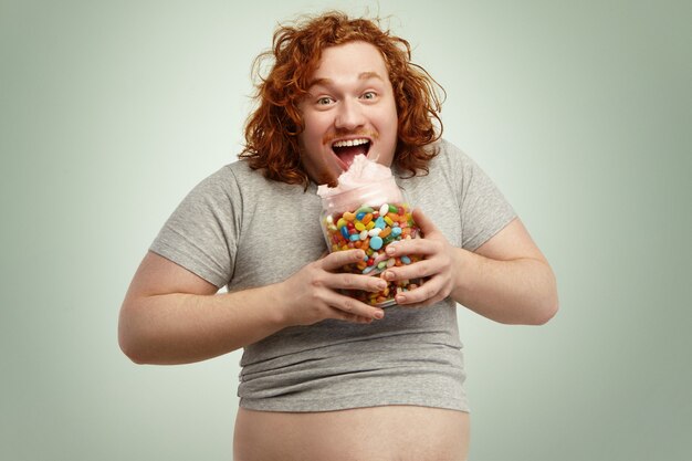 キャンディーフロスを食べながら口を大きく開けて生姜の巻き毛を持つ幸せな肥満の若い男を興奮