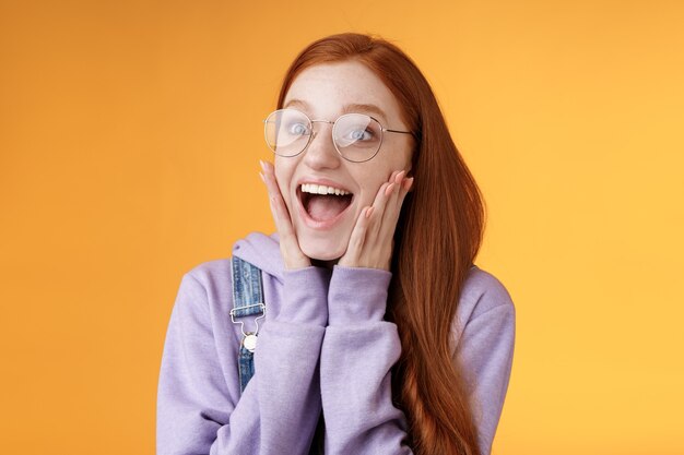 Excited happy smiling broadly redhead girlfriend drop jaw grinning thrilled look left astonished touch cheeks reacting awesome stunning news screaming happiness, standing orange background.