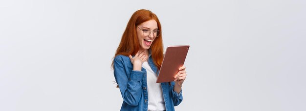 Excited happy and surprised cheerful redhead woman messaging with friend using gadget holding digita