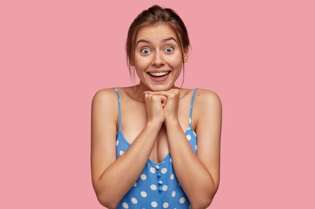 Excited happy surprised European young woman holds chin, dressed in casual blue polka dot dress, looks with eyes full of happiness