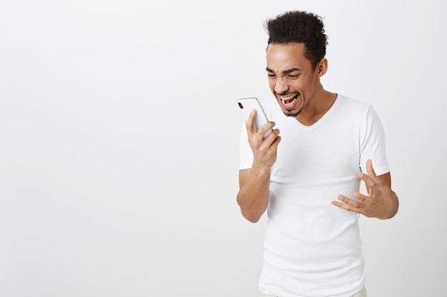 Взволнованный и счастливый торжествующий афроамериканец смотрит на экран мобильного телефона с взволнованным лицом и радуется
