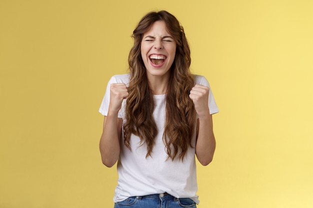 Excited happy triumphing joyful caucasian girl close eyes fist pump celebratory happiness gesture yelling yeah success reach goal achievement dancing victory winning feel relieved yellow background