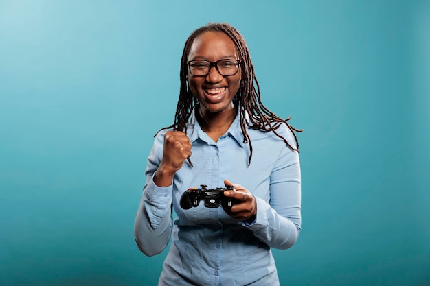 試合の勝利を祝っている間、コンソールでビデオゲームをプレイしている興奮した幸せな女性。青い背景の上に立っている間競争力のあるゲームの勝利を楽しんで幸せな楽しい若い大人の人。 Premium写真