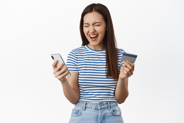 Excited happy woman scream from happiness winning money cashback holding credit card with mobile phone and rejoice standing against white background and laughing