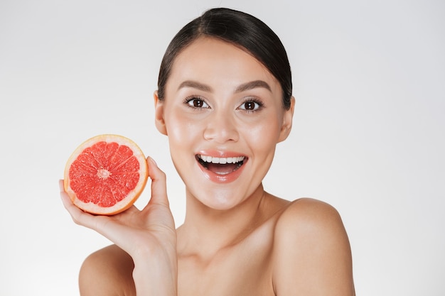 возбужденной счастливой женщины со здоровой свежей кожей, держащей сочный красный грейпфрут и смотрящей на камеру с улыбкой, изолированной над белой
