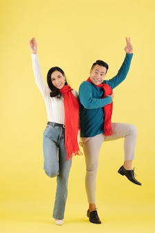 Excited happy young asian couple in wool winter scarf dancing together against yellow background