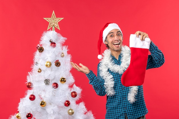 青い縞模様のシャツにサンタクロースの帽子をかぶって、クリスマスツリーの近くにクリスマスの靴下を持って興奮して幸せな若い男