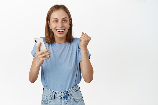 Giovane donna eccitata e felice che tiene smartphone, vince soldi sul cellulare, raggiunge l'obiettivo quotidiano in app, salta dalla felicità e sorride soddisfatto, sfondo bianco