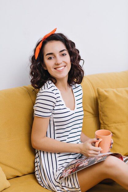 Excited happy young woman resting on couch, reading magazine, having free time in modern apartment. Cheerful mood, drinking tea, enjoying, relax, comfort at home
