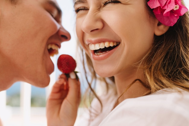 Excited happy young woman with cute pink flower in light-brown hair feeding her laughing husband with fresh strawberry. Close-up portrait of romantic enjoying honeymoon and eating berries