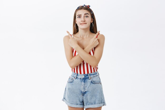 Excited hesitant attractive caucasian woman in striped top and shorts biting lip nervously crossing hands against body pointing in different directions left and right cannot make choice, hesitating