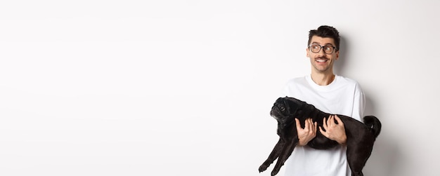 Foto gratuita eccitato ragazzo hipster con il suo cane che guarda a sinistra il logo bel proprietario di animali domestici porta un carlino nero e lo fissa