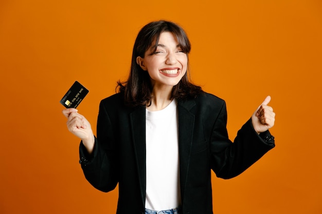 Excited holding card young beautiful female wearing black jacket isolated on orange background