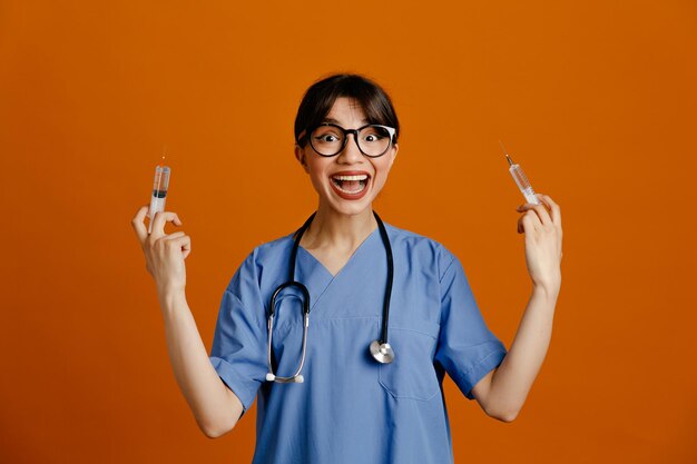 주황색 배경에 격리된 균일한 청진기를 입고 흥분된 주사기 젊은 여성 의사