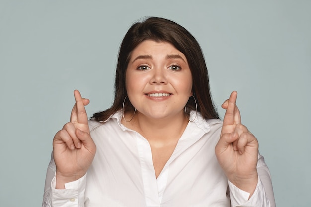 Free photo excited hopeful young plus size female employee in formal shirt crossing fingers for good luck before job interview. overweight brunette student girl hoping for the best, posing with crossed fingers