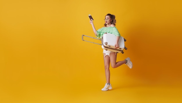 Excited hurry young woman holding suitcase and showing passport on yellow background