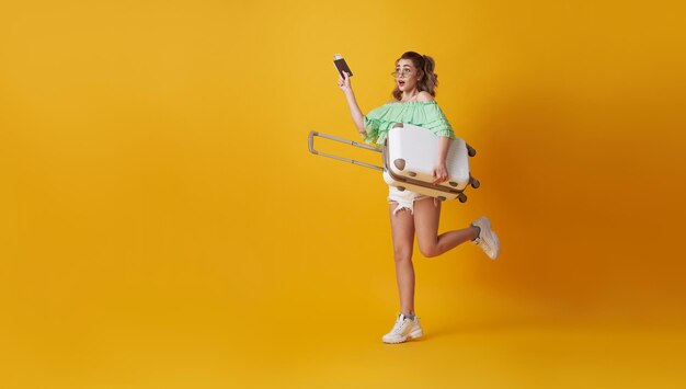 Excited hurry young woman holding suitcase and showing passport on yellow background