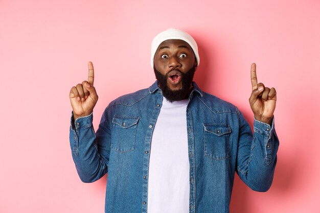 Excited and impressed Black man showing amazing promo, pointing fingers up at logo, standing against pink background