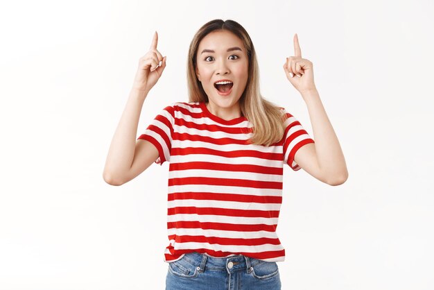 Excited impressed cheerful young asian blond girl happy finally product in stock pointing up index finger amused telling you cool copy space open mouth smiling fascinated standing white background